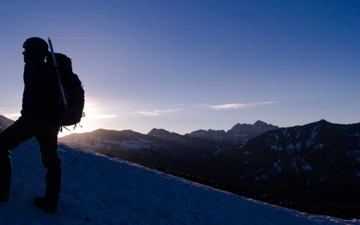 The silhouette of a person wearing a backpack is illuminated by the sun behind them. They appear to be standing on a snowy incline with mountains in the background.